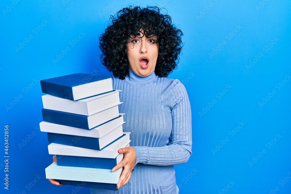 Young middle east woman holding a pile of books afraid and shocked with surprise and amazed expression, fear and excited face.