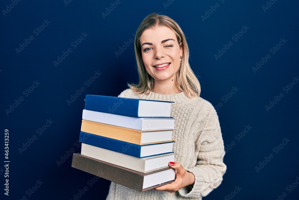 Beautiful caucasian woman holding a pile of books winking looking at the camera with sexy expression, cheerful and happy face. Beautiful caucasian woman holding a pile of books winking looking at the camera with sexy expression, cheerful and happy face.