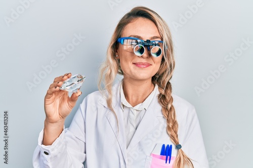 Beautiful young blonde woman holding brilliant diamond stone wearing magnifier glasses smiling looking to the side and staring away thinking.