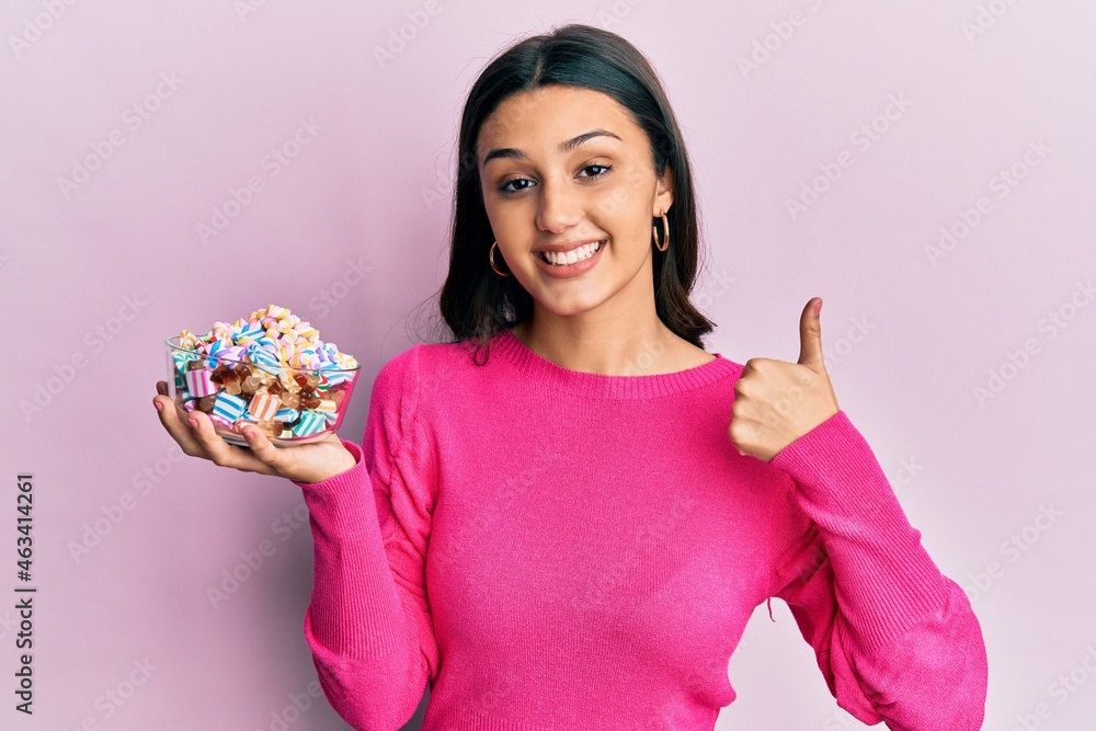 Young hispanic woman holding bowl of sugar candy smiling happy and positive, thumb up doing excellent and approval sign Young hispanic woman holding bowl of sugar candy smiling happy and positive, thumb up doing excellent and approval sign