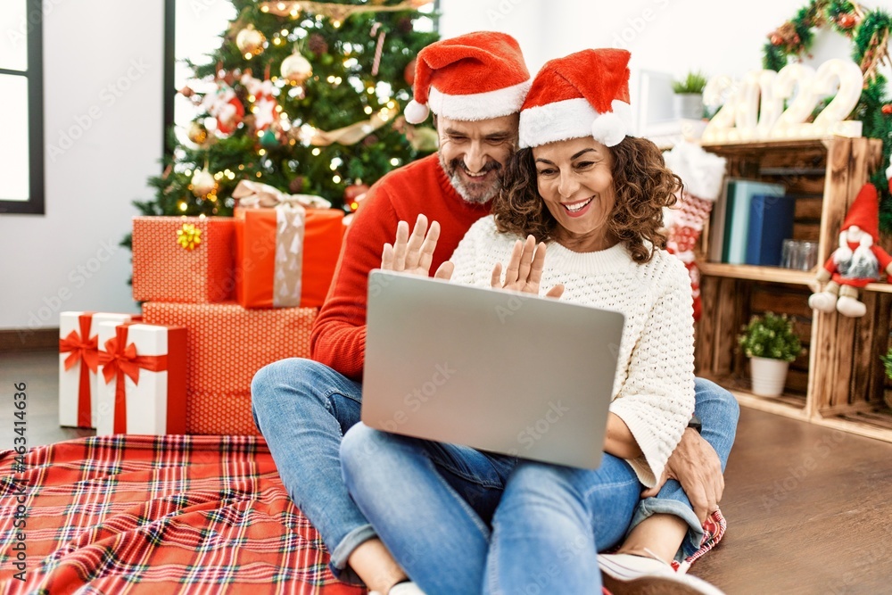 Middle age hispanic couple wearing christmas hat. Sitting on the floor having video call using laptop at home. Middle age hispanic couple wearing christmas hat. Sitting on the floor having video call using laptop at home.