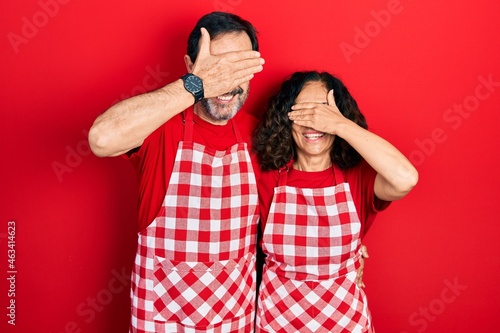 Middle age couple of hispanic woman and man wearing cook apron smiling and laughing with hand on face covering eyes for surprise. blind concept.