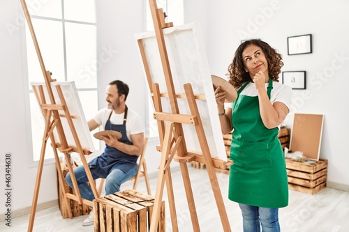Hispanic middle age woman and mature man at art studio serious face thinking about question with hand on chin, thoughtful about confusing idea