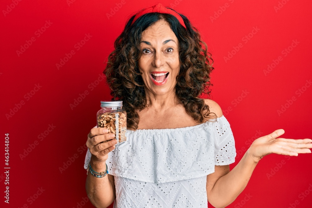 Middle age hispanic woman holding jar with walnuts celebrating achievement with happy smile and winner expression with raised hand Middle age hispanic woman holding jar with walnuts celebrating achievement with happy smile and winner expression with raised hand
