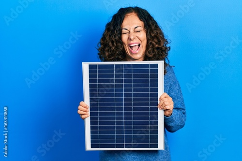 Middle age hispanic woman holding photovoltaic solar panel smiling and laughing hard out loud because funny crazy joke.