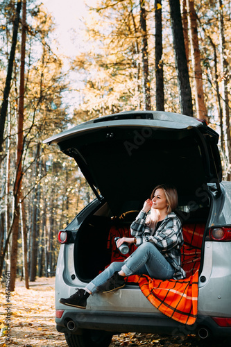 a blonde adult girl in a plaid shirt is drinking coffee sitting in the open trunk of a car. picnic in the car.