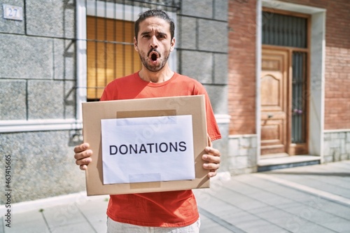 Young hispanic man holding donations box for charity outdoors afraid and shocked with surprise and amazed expression, fear and excited face.