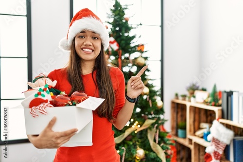 Young brunette woman standing by christmas tree holding decoration pointing aside worried and nervous with forefinger  concerned and surprised expression