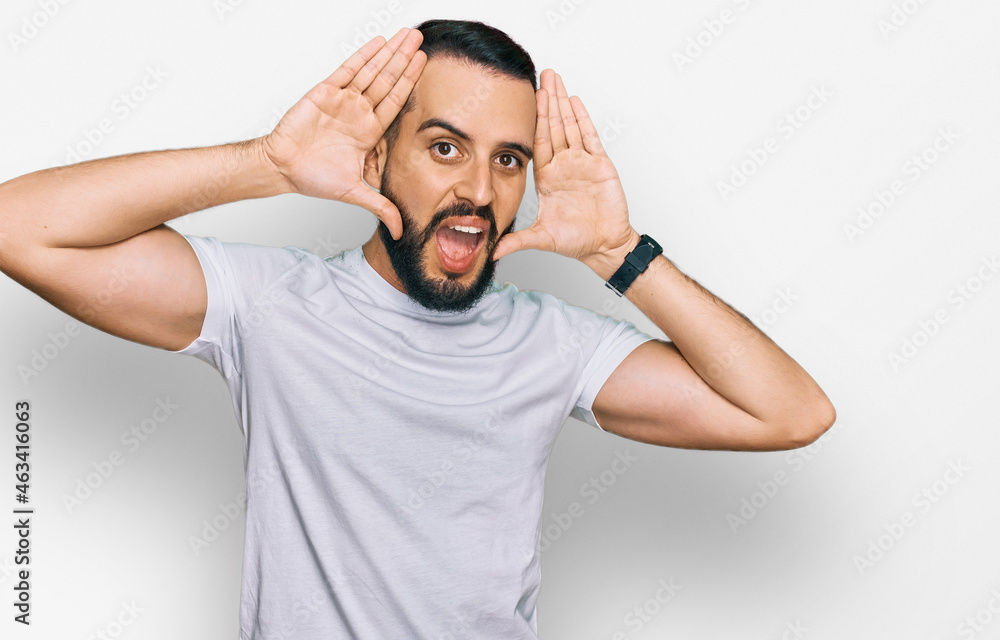 Young man with beard wearing casual white t shirt smiling cheerful playing peek a boo with hands showing face. surprised and exited Young man with beard wearing casual white t shirt smiling cheerful playing peek a boo with hands showing face. surprised and exited
