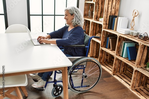 Leinwand Poster Middle age grey-haired disabled woman using laptop sitting on wheelchair at home