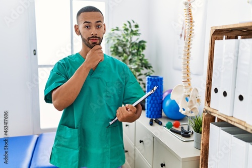 African american physiotherapist man working at pain recovery clinic looking confident at the camera smiling with crossed arms and hand raised on chin. thinking positive.