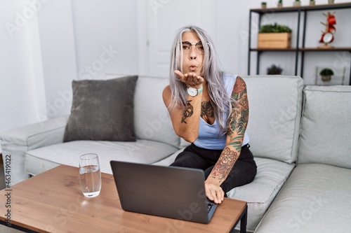 Middle age grey-haired woman using laptop at home looking at the camera blowing a kiss with hand on air being lovely and sexy. love expression.