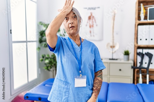 Middle age grey-haired woman wearing physiotherapist uniform at medical clinic surprised with hand on head for mistake, remember error. forgot, bad memory concept.