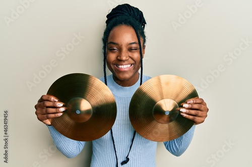 African american woman with braided hair holding golden cymbal plates winking looking at the camera with sexy expression, cheerful and happy face.