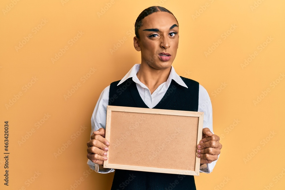 Hispanic man wearing make up and long hair holding empty corkboard clueless and confused expression. doubt concept. Hispanic man wearing make up and long hair holding empty corkboard clueless and confused expression. doubt concept.