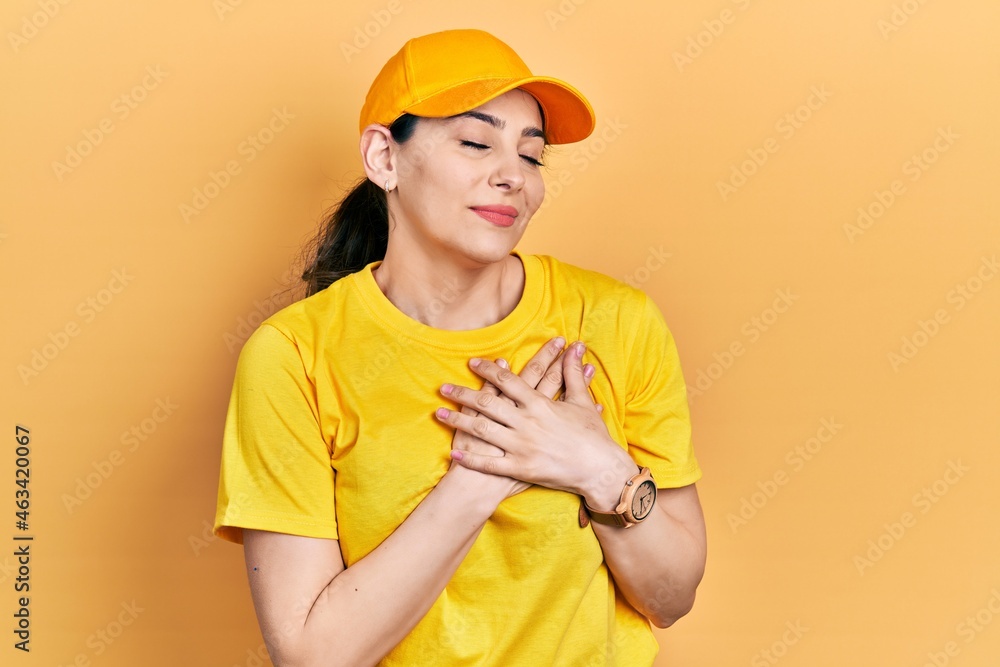 Young hispanic woman wearing delivery uniform and cap smiling with hands on chest with closed eyes and grateful gesture on face. health concept.