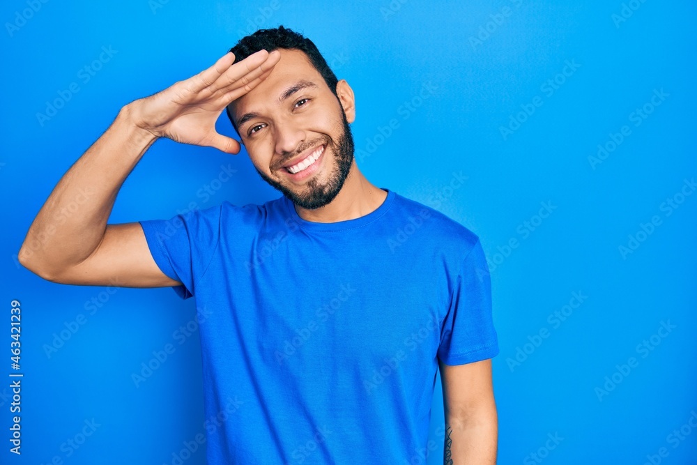 Hispanic man with beard wearing casual blue t shirt very happy and smiling looking far away with hand over head. searching concept. - obrazy, fototapety, plakaty 