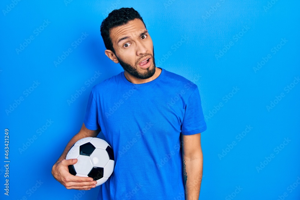 Hispanic man with beard holding soccer ball in shock face, looking skeptical and sarcastic, surprised with open mouth Hispanic man with beard holding soccer ball in shock face, looking skeptical and sarcastic, surprised with open mouth