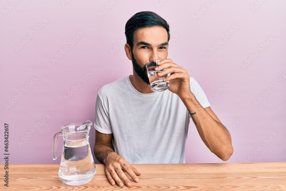 Hispanic man with beard drinking a glass of fresh mineral water sitting on the table