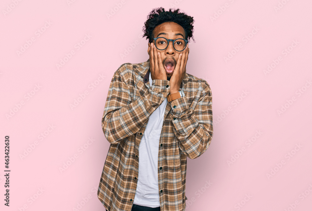 Young african american man with beard wearing casual clothes and glasses afraid and shocked, surprise and amazed expression with hands on face