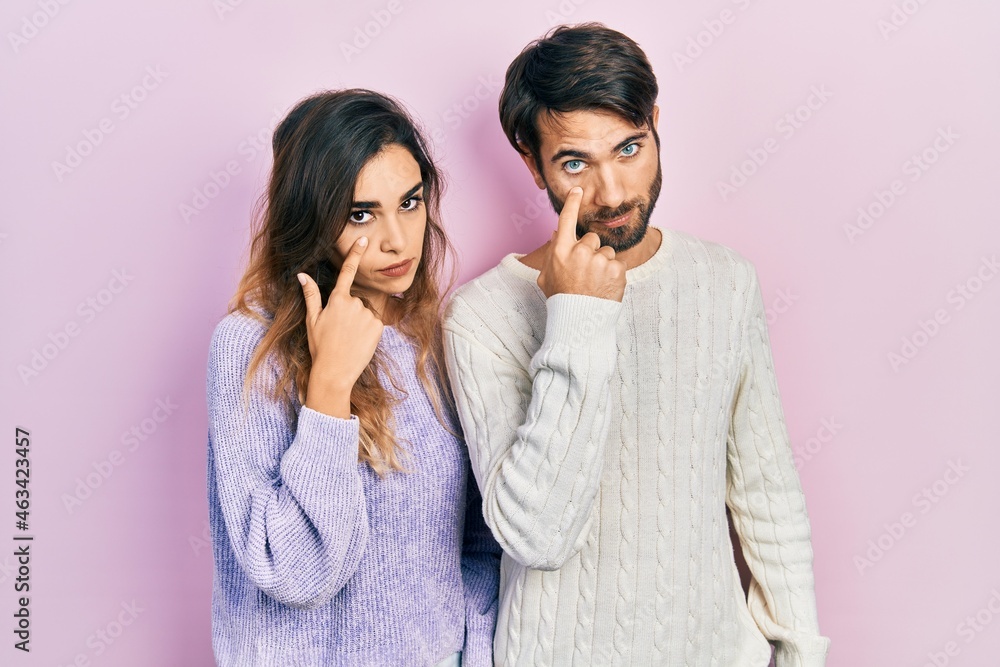 Young hispanic couple wearing casual clothes pointing to the eye watching you gesture, suspicious expression Young hispanic couple wearing casual clothes pointing to the eye watching you gesture, suspicious expression