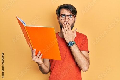 Young hispanic man reading book covering mouth with hand, shocked and afraid for mistake. surprised expression