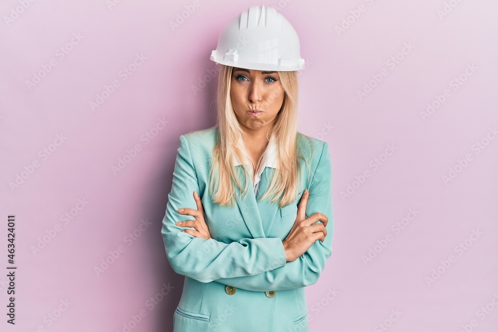 Young blonde woman wearing architect hardhat puffing cheeks with funny face. mouth inflated with air, catching air. Young blonde woman wearing architect hardhat puffing cheeks with funny face. mouth inflated with air, catching air.