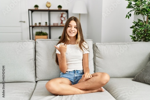 Young brunette teenager sitting on the sofa at home smiling with happy face looking and pointing to the side with thumb up.