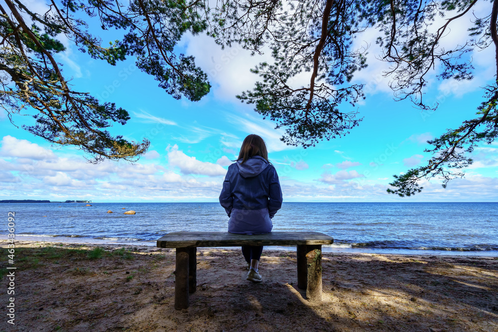 Woman from behind sitting on a bench facing the sea and tree branches around her.