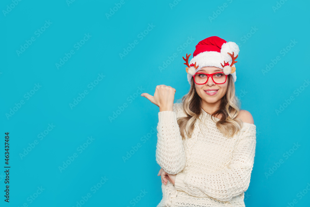 A young charming smiling blonde woman in glasses with deer antlers, a white knitted warm sweater and Christmas Santa hat points at empty copy space for text or design isolated on color blue background A young charming smiling blonde woman in glasses with deer antlers, a white knitted warm sweater and Christmas Santa hat points at empty copy space for text or design isolated on color blue background
