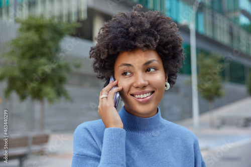 Headshot of pleasant looking young woman enjoys cellphone communication uses contact application talks via modern device wears casual turtleneck poses against urban background. Generation concept