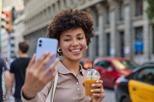Positive curly haired young woman dressed casually drinks smoothie takes selfie records video smiles happily poses at busy road with transport stands against blurred background. Urban lifestyle