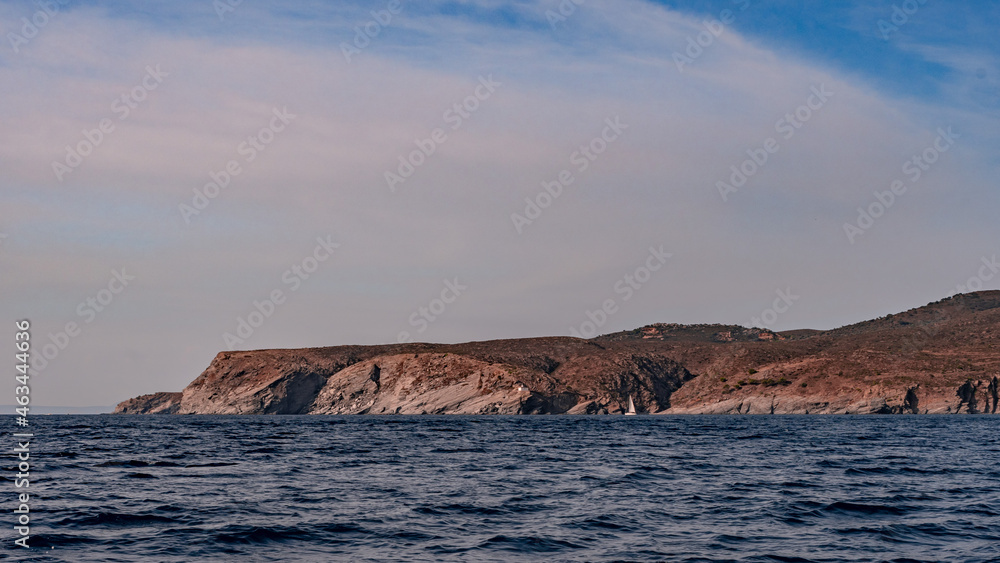 Seaside cliffs and sailboat with Calanans lighthouse next to Cadaques and S'Ocelleta headland, Catalonia, Spain.