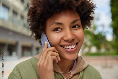 Close up shoot of cheerful woman smiles toothily answers call has pleasant conversation via smartphone chats with friend dressed casually poses against blurred background outdoors. Leisure concept