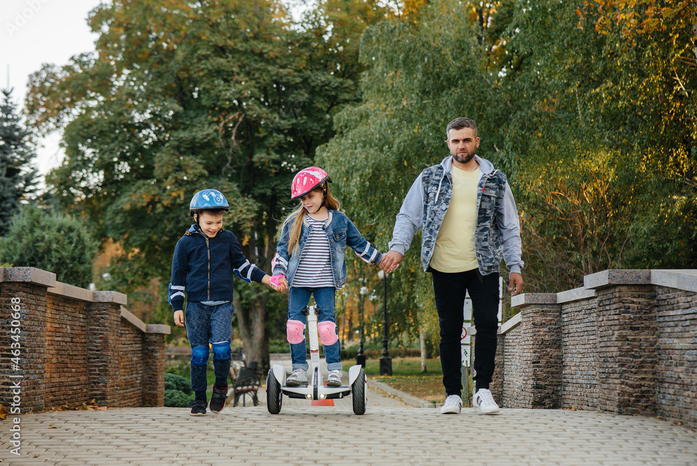 Fototapeta premium A father helps and teaches his young children to ride a Segway in the Park during sunset. Family vacation in the Park.