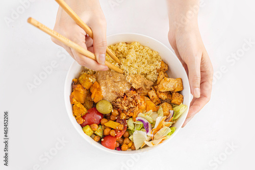 Vegan bowl with couscous, fried tofu, chickpeas, vegetables and peanut sauce in woman's hands with foodsticks. White background, topview. photo