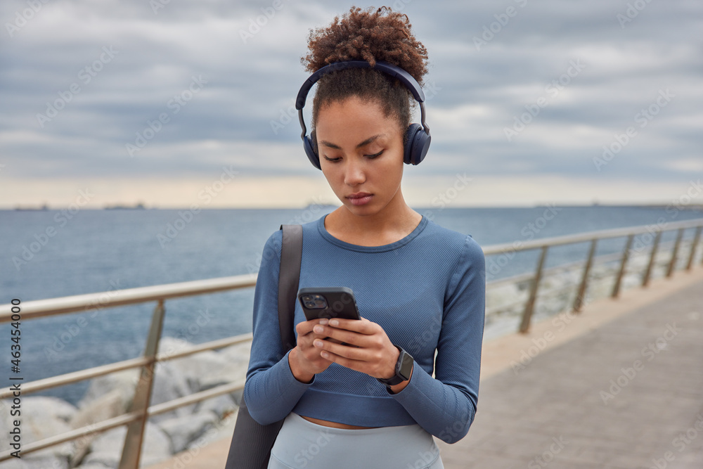Outdoor shot of sporty woman dressed in spotswear checks music from playlist holds mobile phone listens audio track in headphones carries karemat strolls along sea against cloudless blue sky