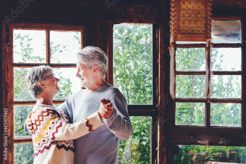 Happy senior couple smile and dance at home enjoying love and relationship together. Active old man and woman have fun in indoor leisure activity. Nature woods view from windows