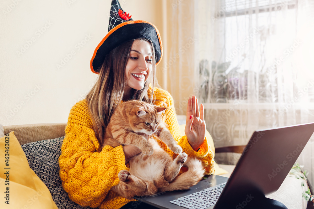 Halloween lockdown. Woman in witch hat holding cat waving hi in video chat on laptop. Holidays during coronavirus - obrazy, fototapety, plakaty 