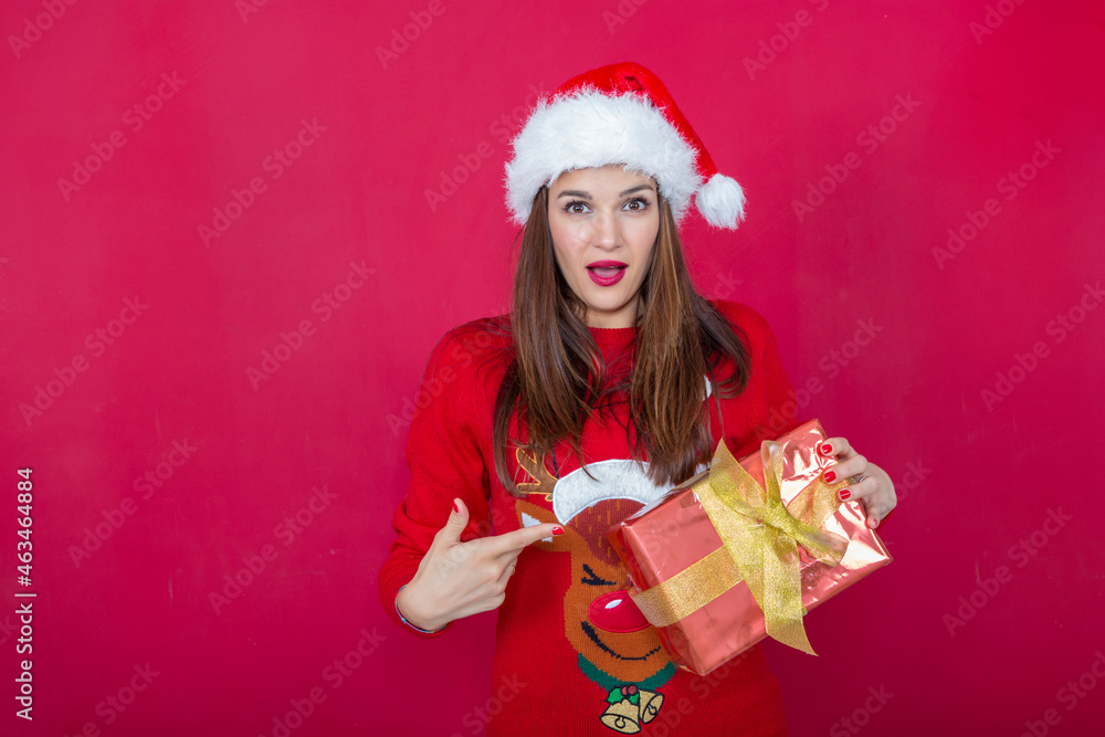 woman in a Santa hat and reindeer sweater, pointing to a gift in her hand, with a surprised gesture. woman in a Santa hat and reindeer sweater, pointing to a gift in her hand, with a surprised gesture.