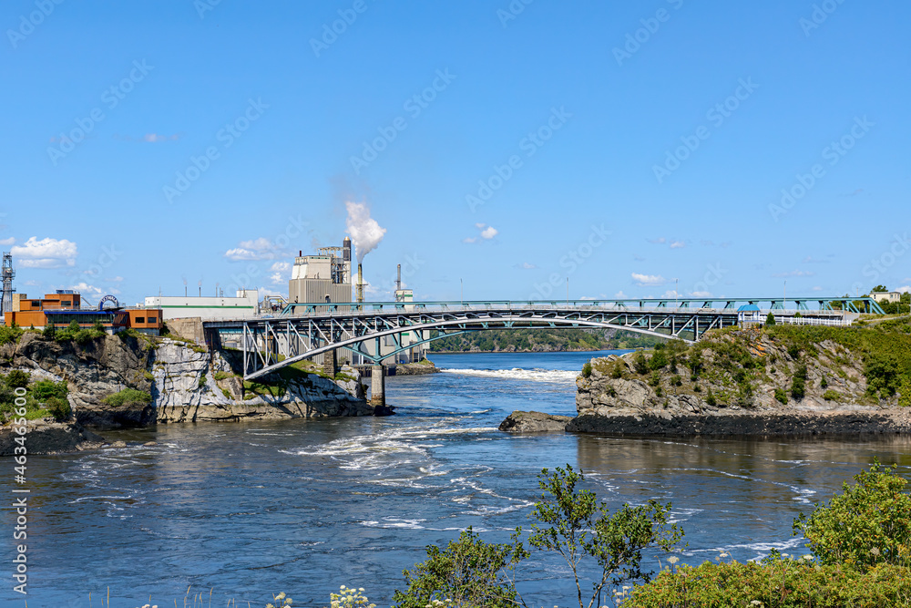 Reversing Falls bridge in saint John, NB, Canada seen from across the river. Pulp mill behind bridge. Blue sky and blue river on a sunny day.