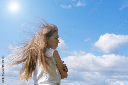 Sunlit young long blond haired woman standing on bright sunlight and blue sky background.Female is hugging and embrtacing herself as concept of calmness harmony and tranquility. Love mood metaphor