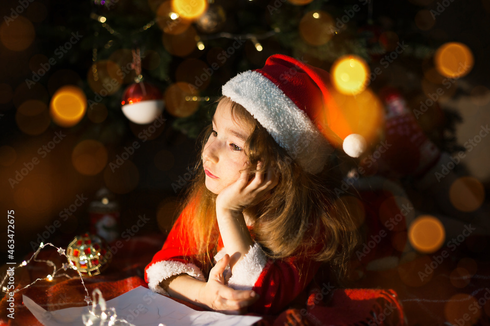 Little girl in a Santa hat and red dress under Christmas tree is dreaming, waiting for the holiday, lying on a plaid blanket. A letter on piece of paper, gifts. New year, Christmas. defocus lights Little girl in a Santa hat and red dress under Christmas tree is dreaming, waiting for the holiday, lying on a plaid blanket. A letter on piece of paper, gifts. New year, Christmas. defocus lights
