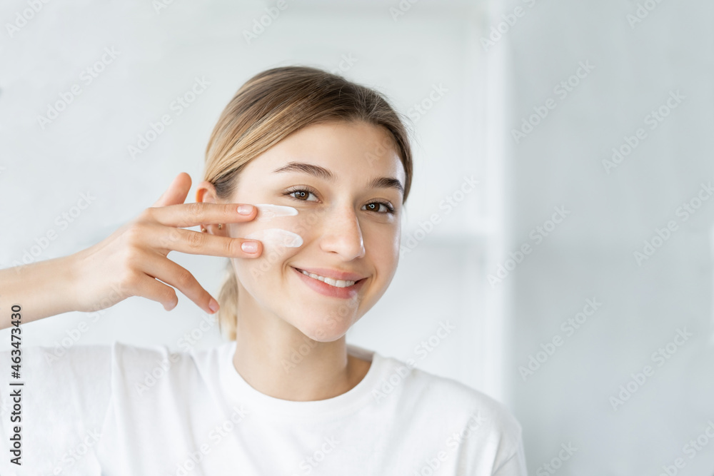 Home skincare. Moisturizing cosmetology. Facial treatment. Cheerful smiling woman applying organic cream on soft glowing face skin at light bathroom. Home skincare. Moisturizing cosmetology. Facial treatment. Cheerful smiling woman applying organic cream on soft glowing face skin at light bathroom.