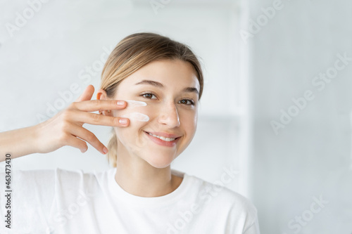 Home skincare. Moisturizing cosmetology. Facial treatment. Cheerful smiling woman applying organic cream on soft glowing face skin at light bathroom.