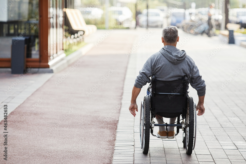 Rear view of mature man with disability riding in wheelchair along the street Rear view of mature man with disability riding in wheelchair along the street