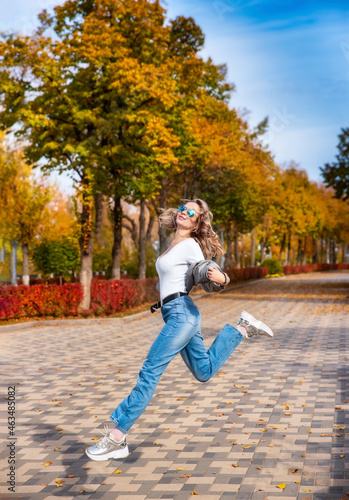 Young happy girl runs along the alley of the autumn park. Carefree energetic girl jumping with happiness. A young woman is enjoying the freedom and good autumn weather. 