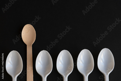 Wooden disposable spoon made from birch tree rises above plastic ones lying on black background. Close-up top view. Copy space for your text. Biodegradable cutlery. Environmental protection theme.