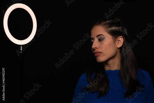 Latin woman. model, young with light skin, tanned. model. with continuous light, shadows and ring of light. Model with eye makeup. green eyes. on black background. dressed in blue. portrait, model.