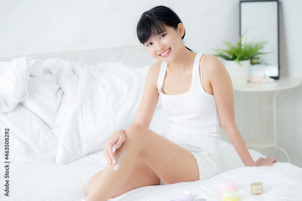 Beautiful young asian woman applying cream or lotion on skin legs with moisturizer having cartridge on bed, beauty asia girl applying skincare touch legs with cosmetic makeup, skin care concept.